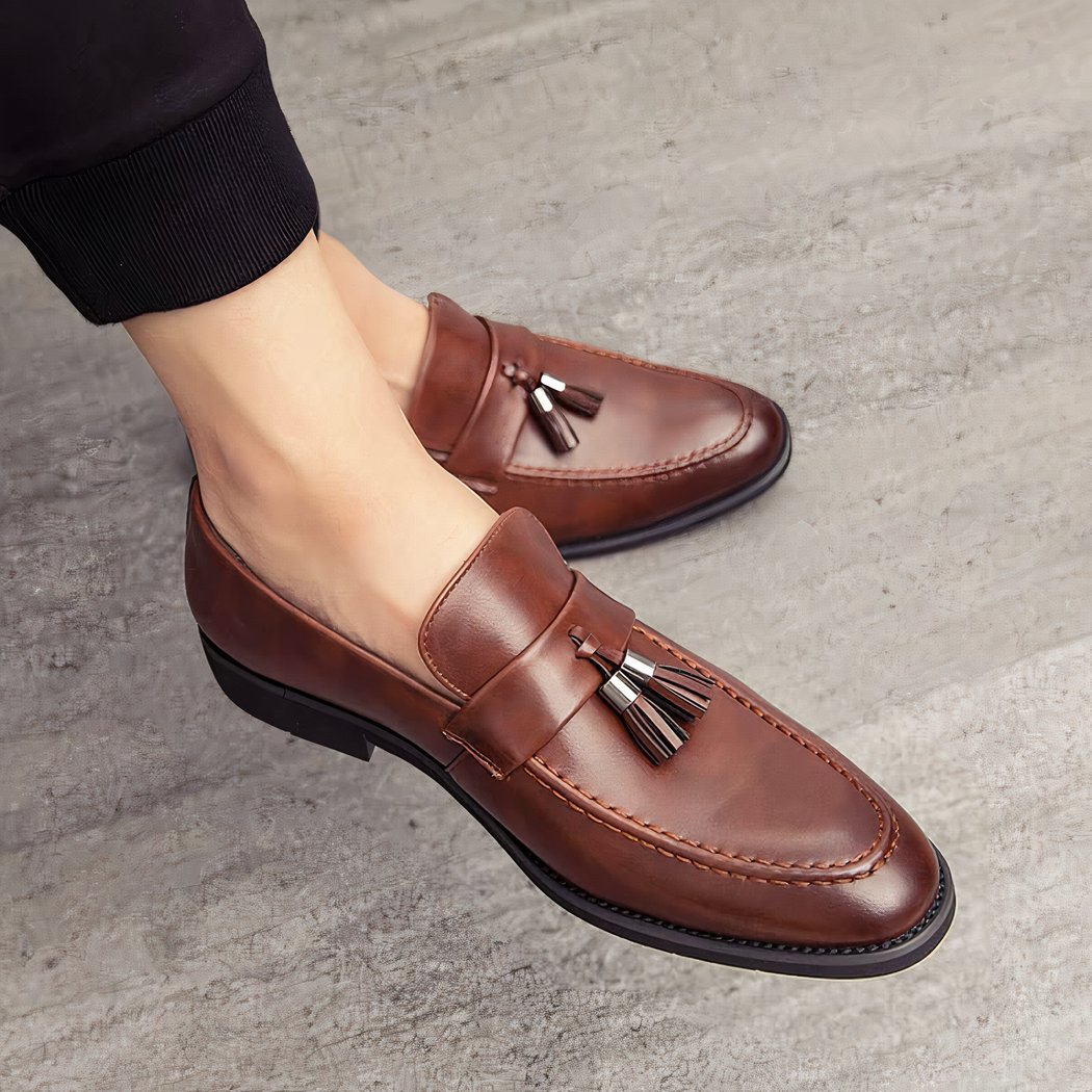 Brown leather loafers with pointed toes and decorative tassels on the feet of a man wearing black pants and crossed legs.