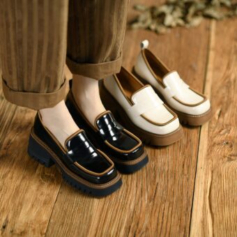 Two pairs of contrasting patent leather loafers, one beige and brown, the other black and brown, at the feet of a woman standing on a parquet floor and wearing wide brown striped pants.