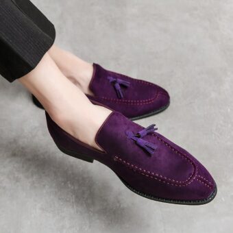 Purple suede loafers with decorative tassels, at the feet of a cross-legged man wearing black suit pants with thin white stripes.