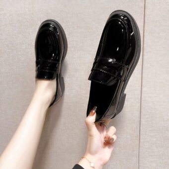Black patent leather loafers, one worn, the other hand-held, on gray background