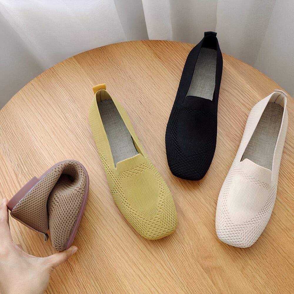 On a wooden table, 3 square-toed moccasins in yellow, black and beige mesh. A hand is seen folding a moccasin to show its suppleness