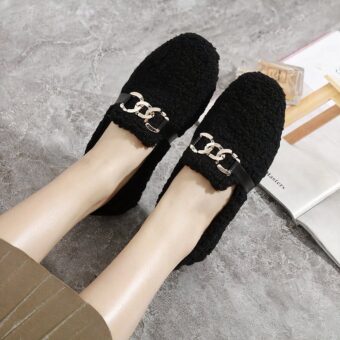 Soft winter moccasins in black bouclette with gold-ringed bits worn on a gray floor with a skirt next to an open book