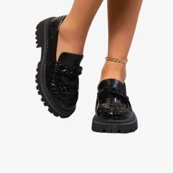 Pair of Black crocodile-look patent loafers for women, worn by a woman whose ankles are visible only. She is wearing an anklet.